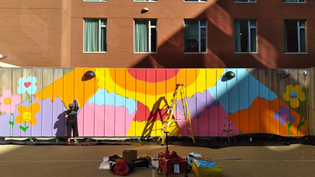 A large work in progress mural on a wood fence in front of a brick building with windows. Amanda paints at the left of fence.