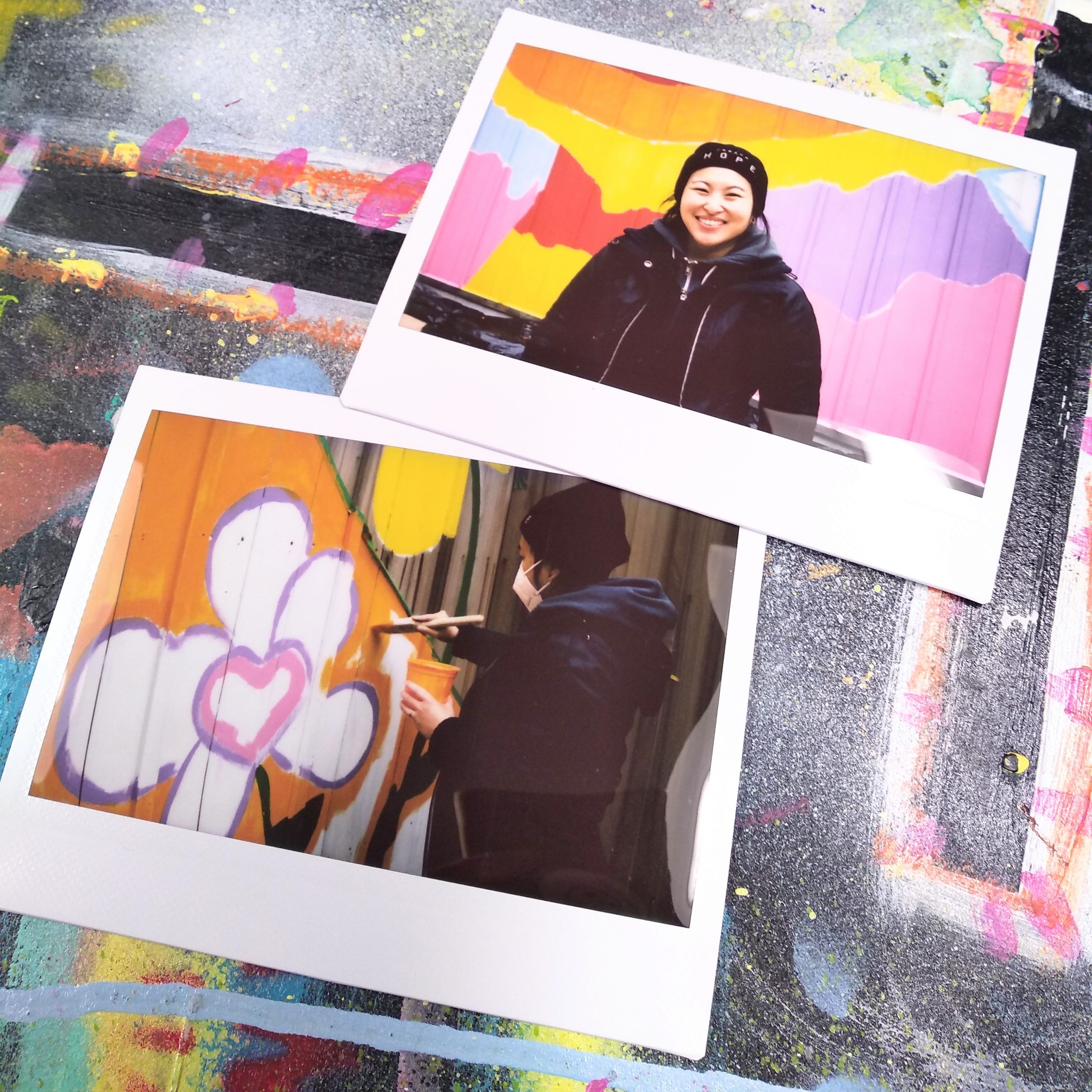 Two Polaroid pictures on colour sprayed surface. Photos of Amanda painting in front of colourful fence.