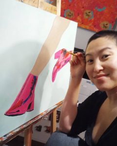 Amanda smiles at the camera as they paint a pair of legs in pink metallic boots. Amanda is in a black tshirt and pants with short hair.